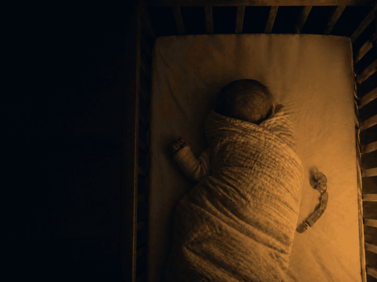 Overhead nursery view of an infant in a crib, gentle amber lamplight, illustrating the difference between back-sleep and prone position.