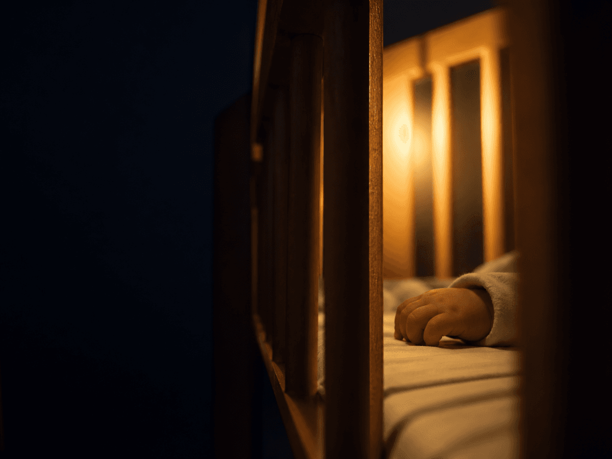 Close-up of wooden crib slats from inside the crib with soft amber backlight and a small infant hand visible, evoking the moment a limb might slip between slats.
